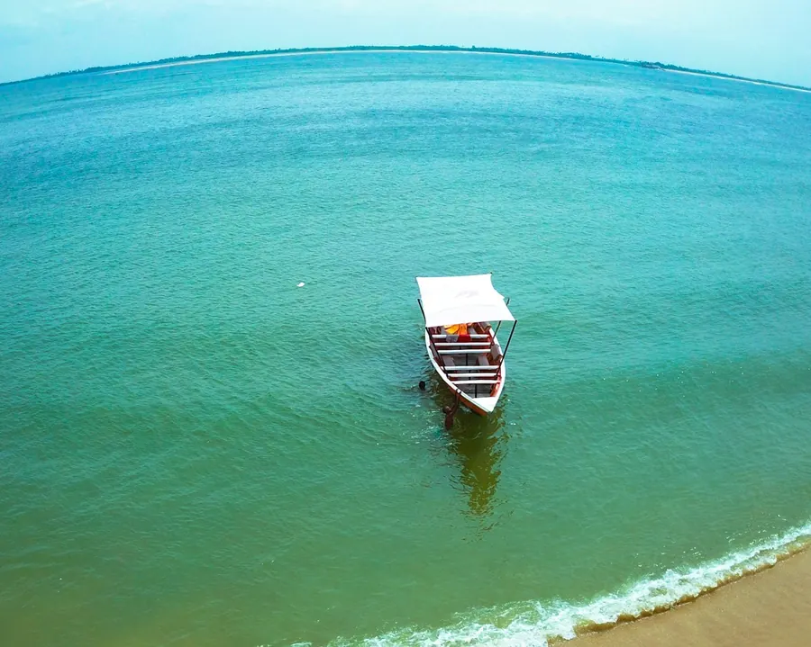 A small covered boat on the green water of the Volta estuary, with a sandy shore