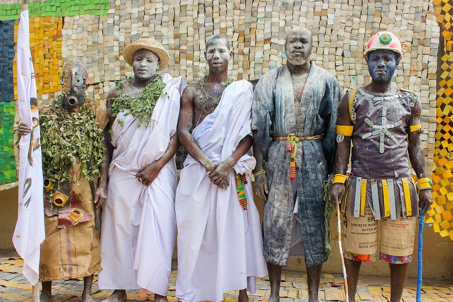 Performers with painted faces in white cloth and costumes standing before a wall of cut yellow gallons at the Afrogallonism exhibition in Accra