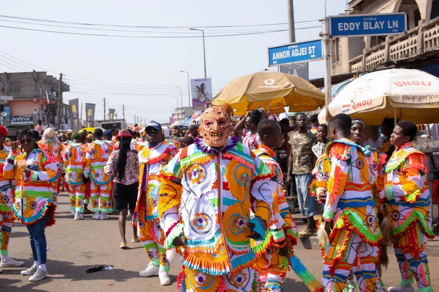 Masqueraders in bright patterned costumes and masks on a busy Takoradi street