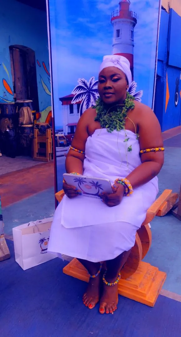 A Ga woman in white traditional dress and beads, seated on a stool, at Homowo
