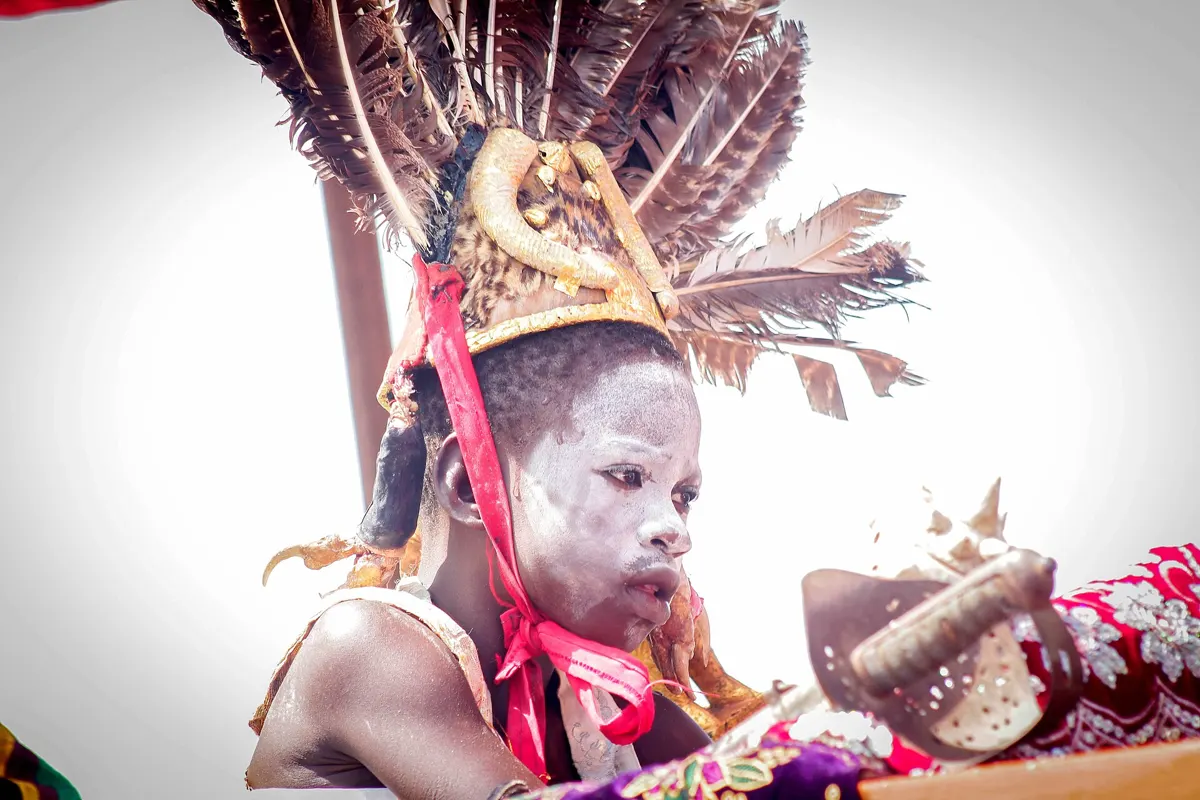 A boy in a feathered headdress and white face paint seated in a palanquin